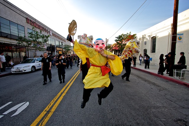 A man in a big head buddha costume leads a crowd down a street in Chinatown.